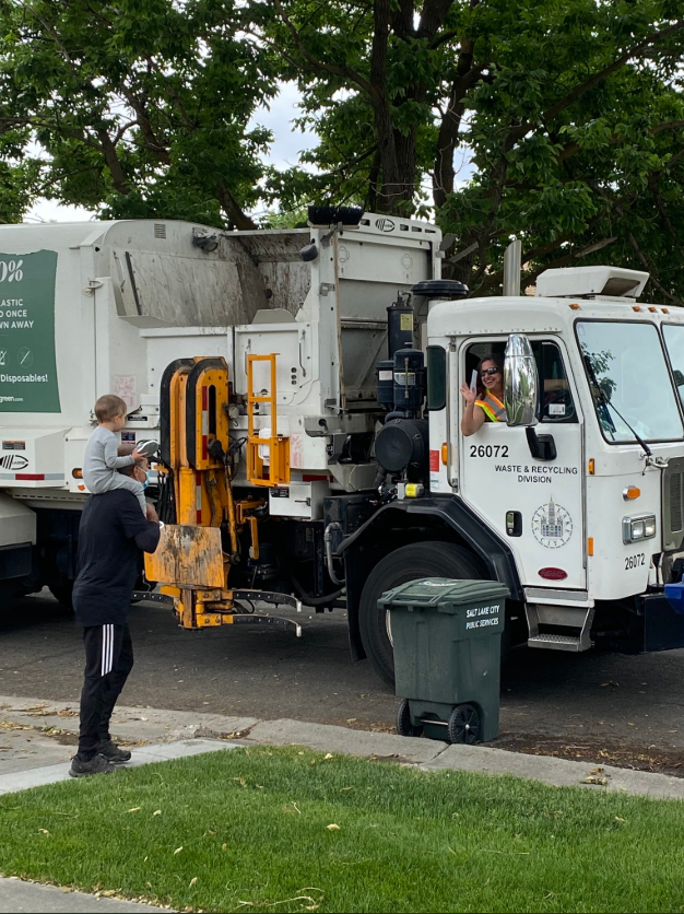 Photo of child sitting on parent's shoulders to wave at Waste & Recycling crew member who is collecting refuse in one of the Salt Lake City garbage trucks. The truck drive is waving back at the child.