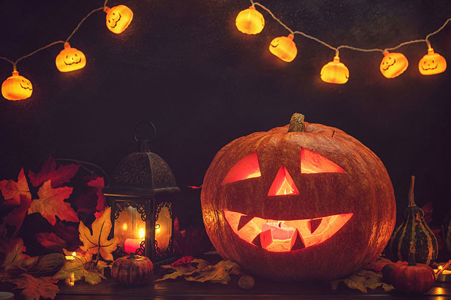 Image of a smiling jack o' lantern in front of a lantern. Leaves are scattered around the pumpkin, and a string of cheerful pumpkin shaped lights frames the top of the image.
