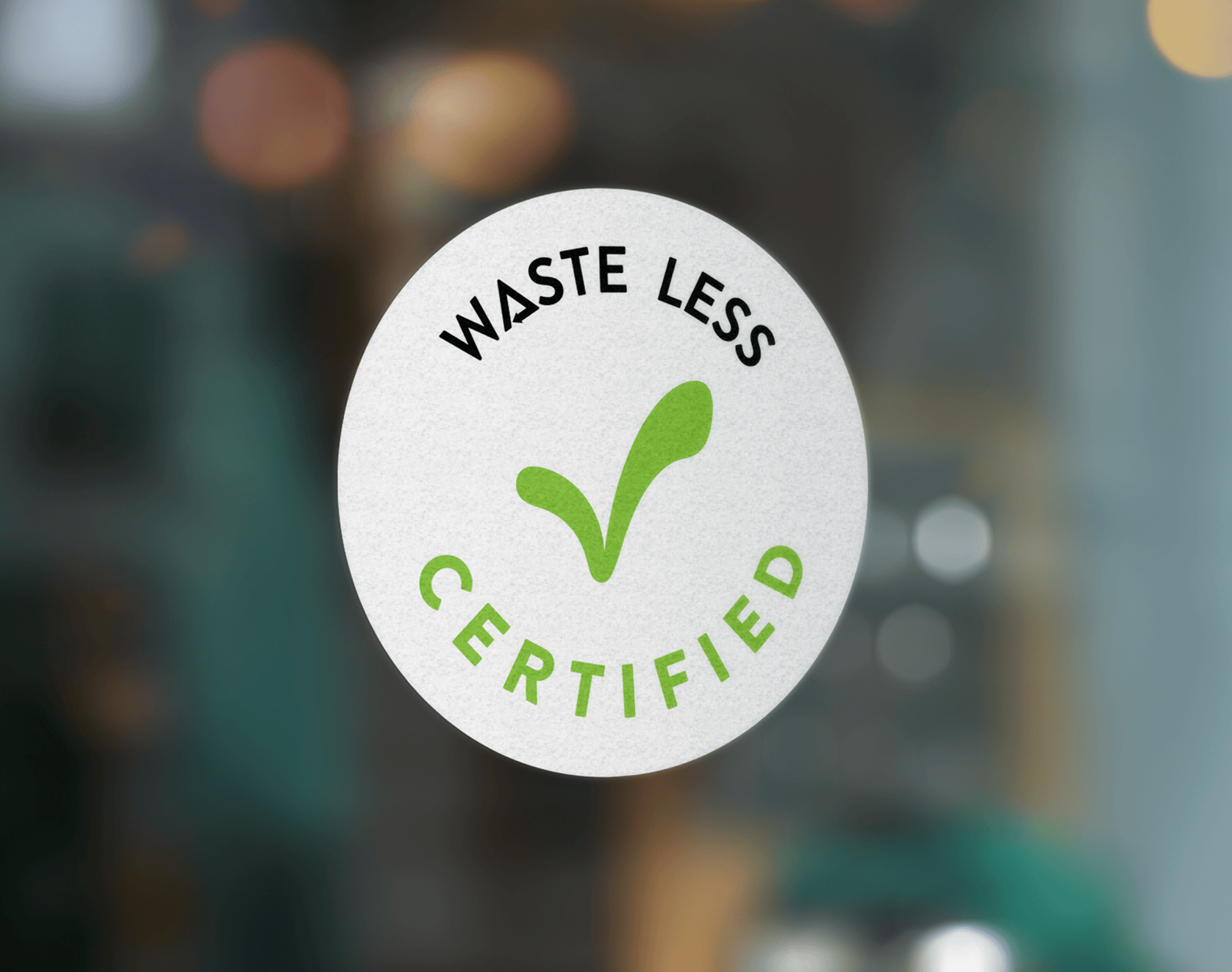 Photo of circular white window decal with green check mark in the center. Text around the edge of the decal reads "Waste Less Certified." 