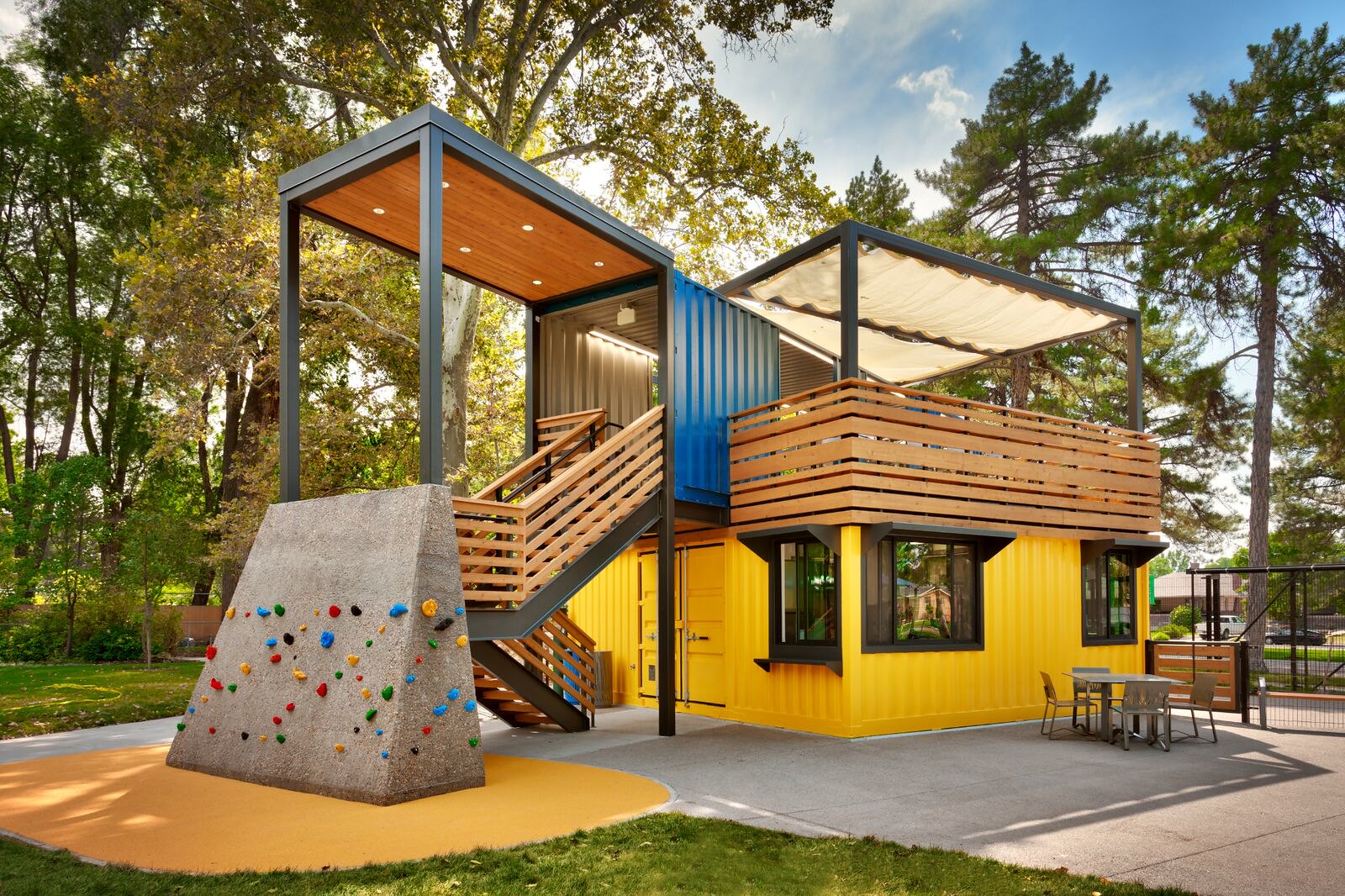 Photo of Tracy Aviary's Bird Feeder Café. The building is constructed out of yellow and blue shipping containers. There is a small rock climbing wall in front for children. Pine and cottonwood trees surround the building.  