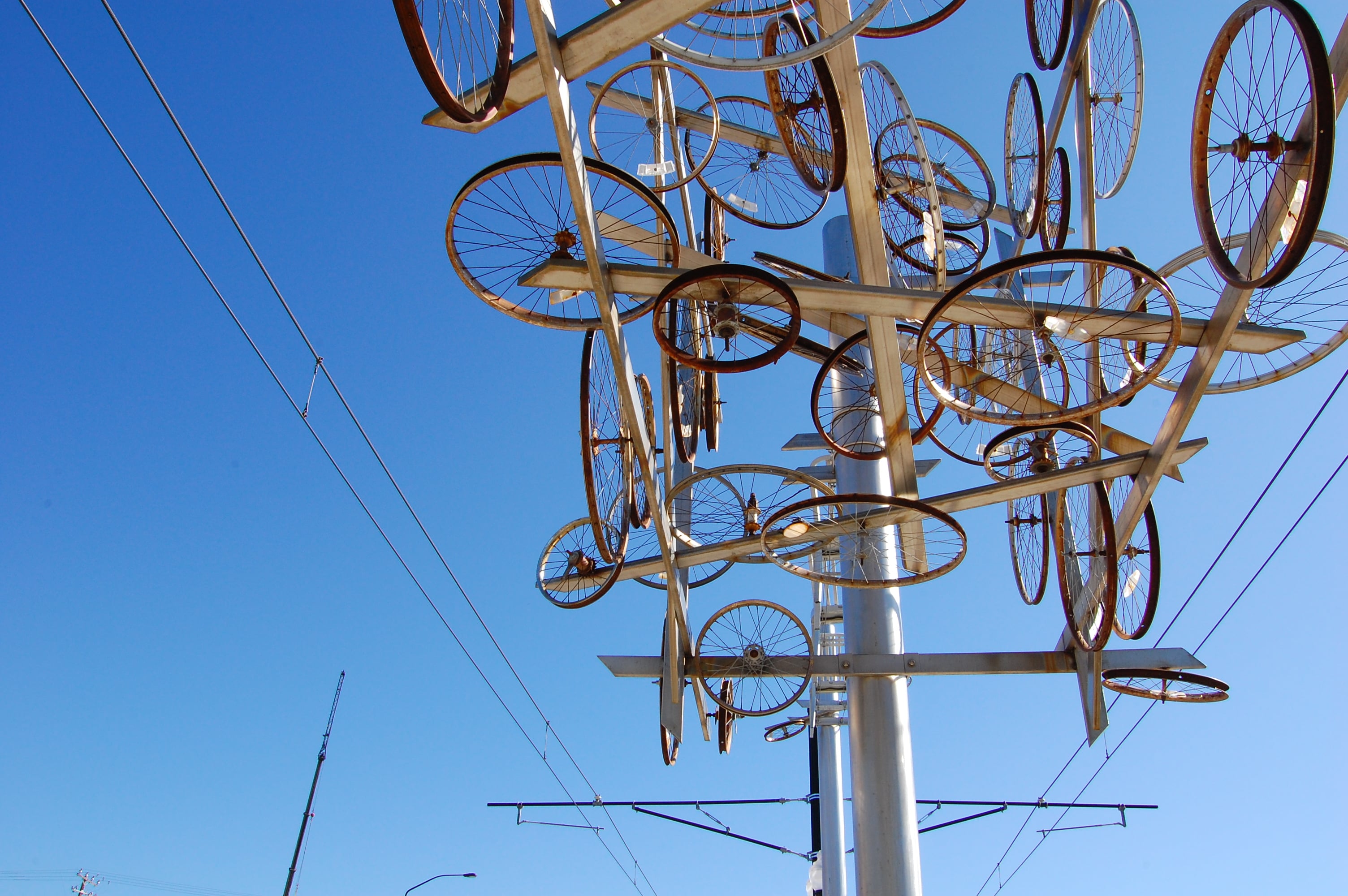 Photo of public art at Salt Lake City TRAX station. The Artwork is a steel poll decorated with different sized bike wheels creating a nest of wheels and steel bars juxtaposed with the steel lines of the light rail and the blue sky. 