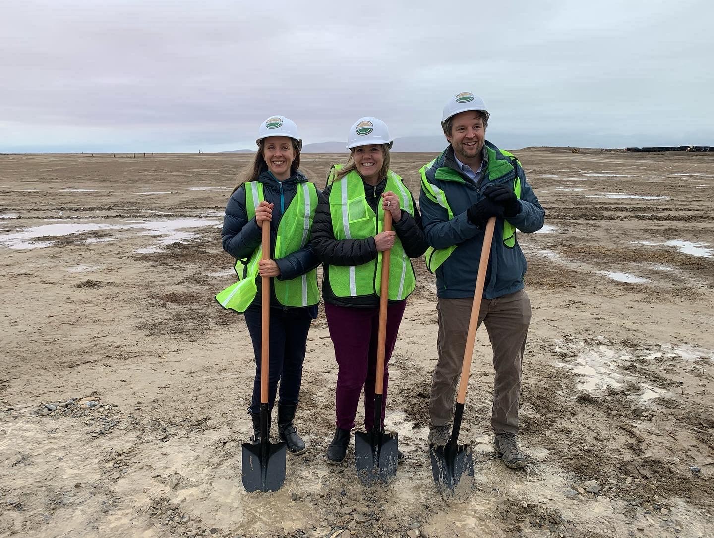 Salt Lake City Sustainability Department's Sophia Nicholas, Debbie Lyons, and Christopher Thomas stand at the groundbreaking event wearing large green reflective vests, white hardhats, and holding large shovels.