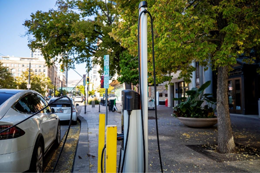 Electric car at a charging station on a city street with trees and buildings in the background.