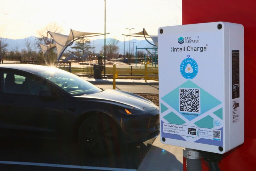 A dark electric car is parked next to a white IntelliCharge charging station mounted on a red pole. In the background is Glendale Park on a sunny day with playground equipment, shade coverings, and trees.
